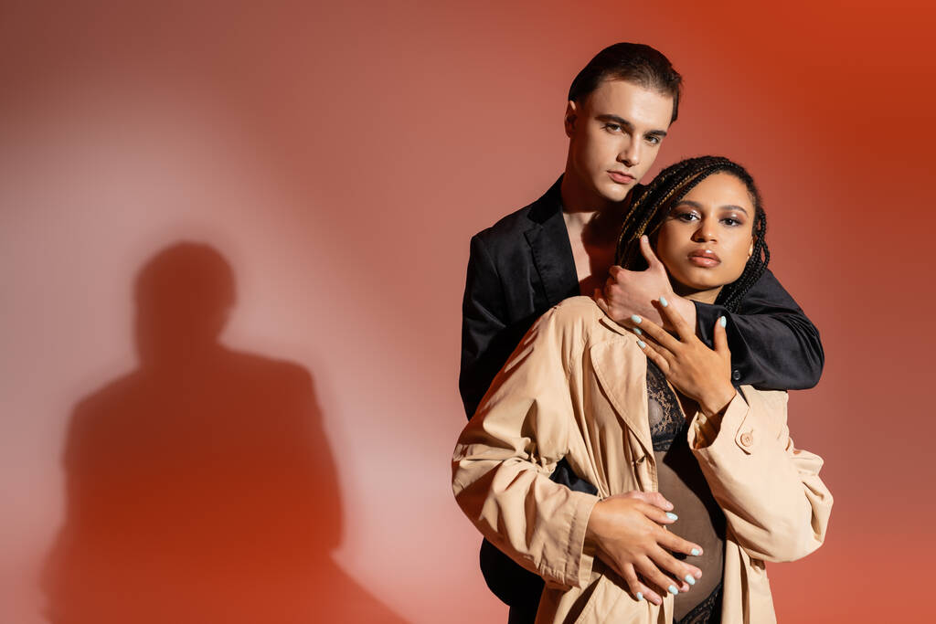 couple interracial sexy, homme élégant en blazer noir et femme afro-américaine en costume de dentelle et trench coat beige regardant la caméra sur fond rouge et rose - Photo, image