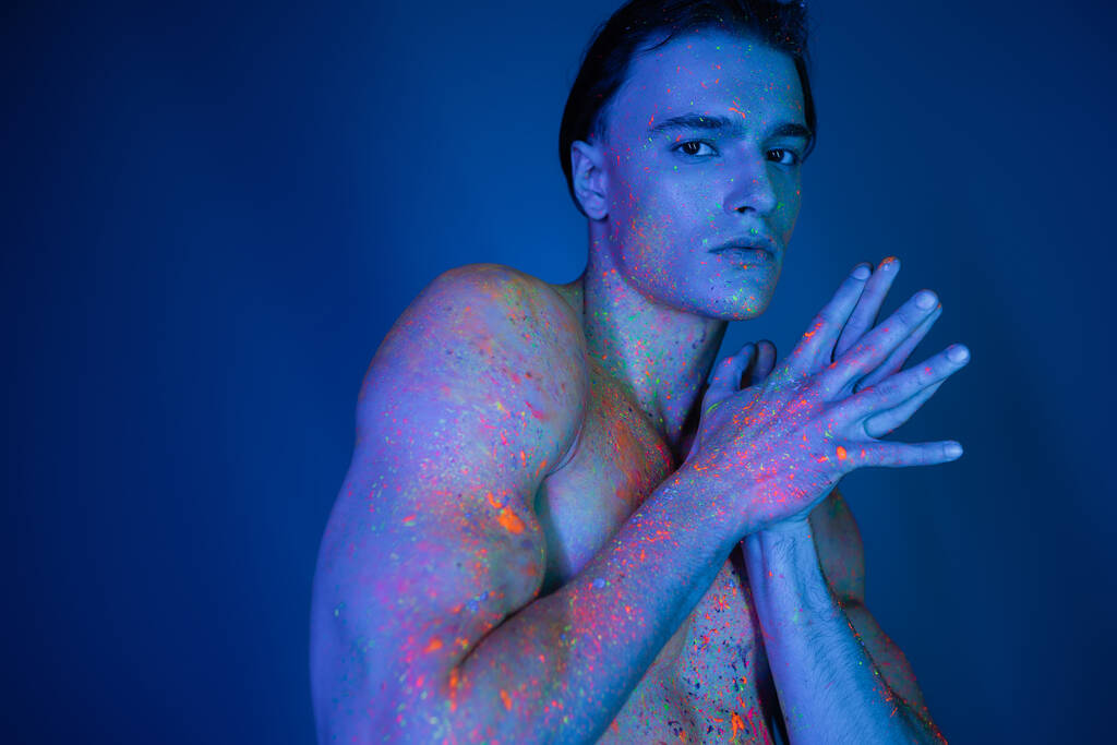 charismatický a sebejistý muž bez trička v zářivém a pestrobarevném neonovém nátěru na tělo, který stojí se spojenýma rukama a dívá se na kameru na modrém pozadí s azurovým efektem osvětlení - Fotografie, Obrázek