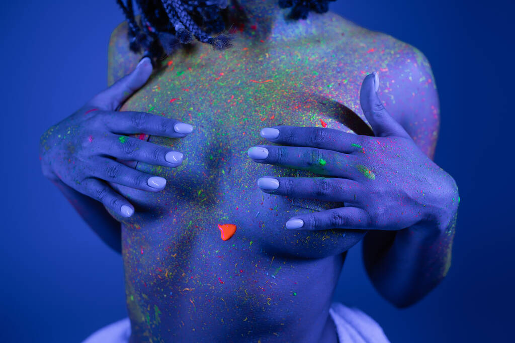 Kısmen genç ve çıplak göğüslü Afro-Amerikan kadını canlı ve renkli vücut boyası ile göğüslerini mavi arka planda ve siyan ışıklandırma efektiyle boyuyor. - Fotoğraf, Görsel