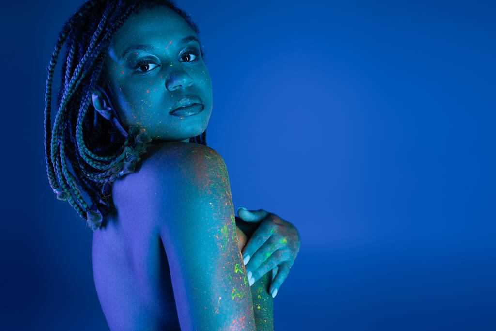 tempting african american woman with dreadlocks covering breast with hands and looking at camera while posing in colorful neon body paint on blue background with cyan lighting effect - Photo, Image