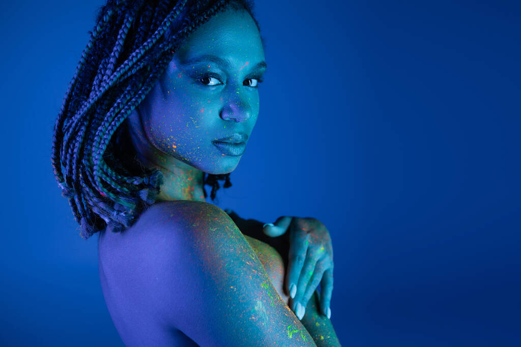 intrigante femme afro-américaine en peinture au néon multicolore sur le corps en regardant la caméra et en couvrant la poitrine avec les mains tout en posant sur fond bleu avec un effet d'éclairage cyan - Photo, image