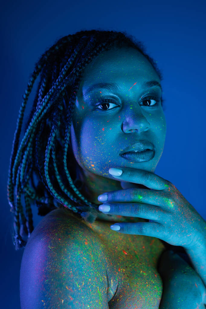 retrato de jovem e atraente mulher afro-americana em pintura de corpo de néon multicolorido segurando a mão perto do rosto e olhando para a câmera no fundo azul com efeito de iluminação ciano - Foto, Imagem