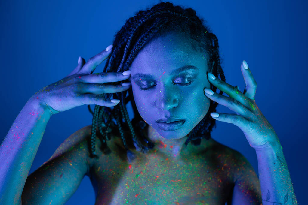 verleidelijk en blote borst Afrikaans amerikaanse vrouw in kleurrijke neon body paint poseren met gesloten ogen en handen in de buurt van gezicht op blauwe achtergrond met cyaan lichteffect - Foto, afbeelding
