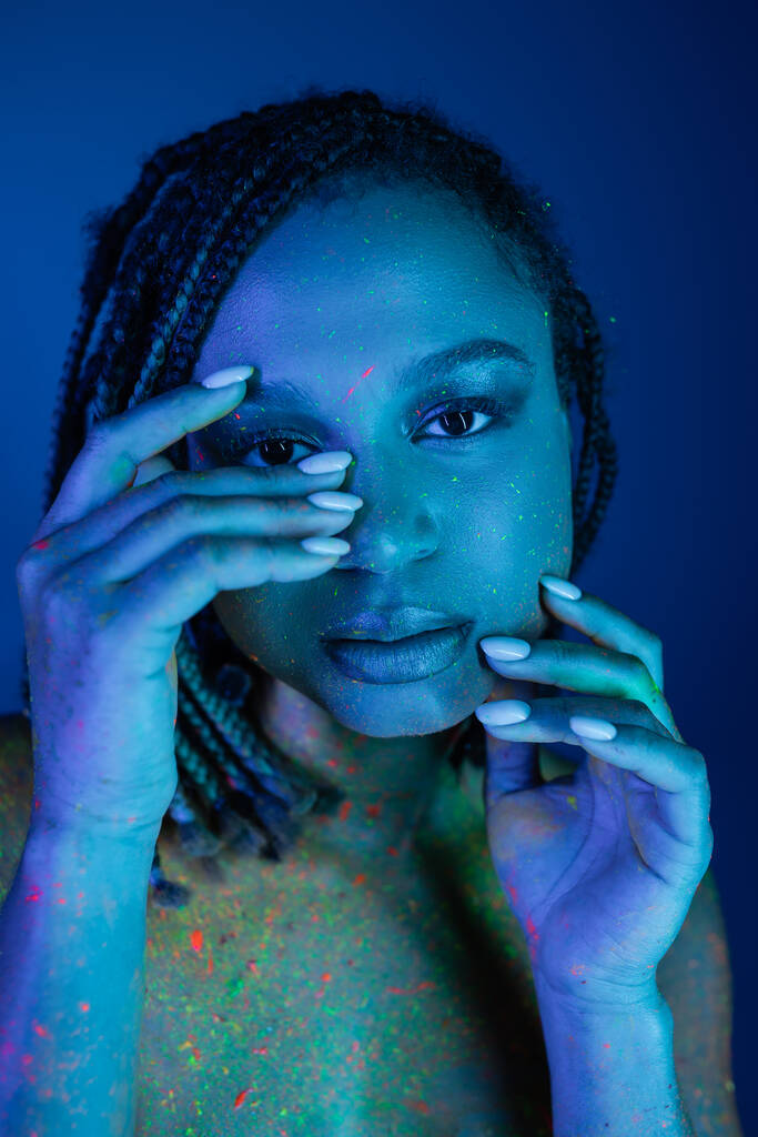 retrato de mujer afroamericana joven y seductora con rastas cogidas de la mano cerca de la cara mientras posa en colorida pintura de cuerpo de neón sobre fondo azul con efecto de iluminación cian - Foto, Imagen
