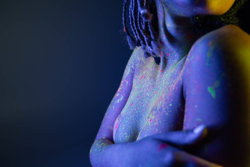 Teilansicht der jugendlichen und nackten afrikanisch-amerikanischen Frau mit Dreadlocks, die in bunter Neon-Körperfarbe posiert und die Brust mit Händen auf blauem Hintergrund mit gelbem Lichteffekt bedeckt - Foto, Bild