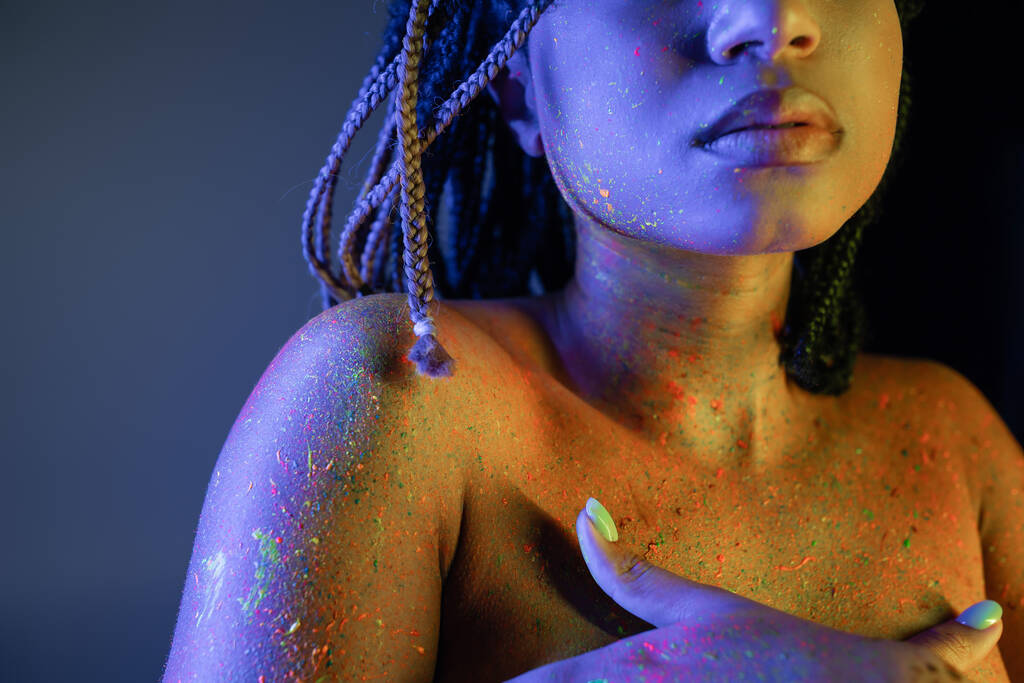 részleges kilátás a fiatal és csupasz mellű afro-amerikai nő sugárzó és színes neon testfesték borító mell kezek kék háttér sárga fényhatás - Fotó, kép