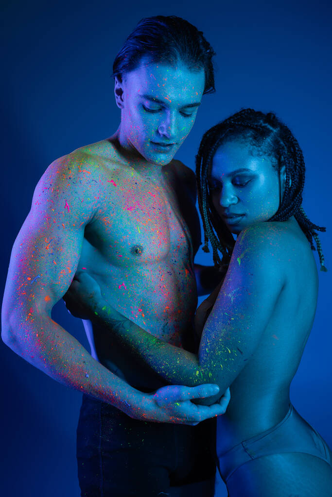 intimer Moment eines gemischtrassigen Paares in bunter Neon-Körperfarbe, das sich auf blauem Hintergrund mit Cyanbeleuchtung umarmt, Mann mit muskulösem Oberkörper und hemdlose Afroamerikanerin mit Dreadlocks - Foto, Bild