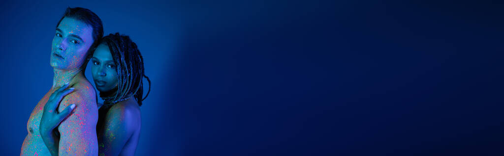 romantisches Paar in bunter Neon-Körperbemalung mit Blick in die Kamera auf blauem Hintergrund mit Cyanbeleuchtung, afrikanisch-amerikanische Frau mit Dreadlocks umarmt sexy hemdlosen Mann mit muskulösem Oberkörper, Banner - Foto, Bild