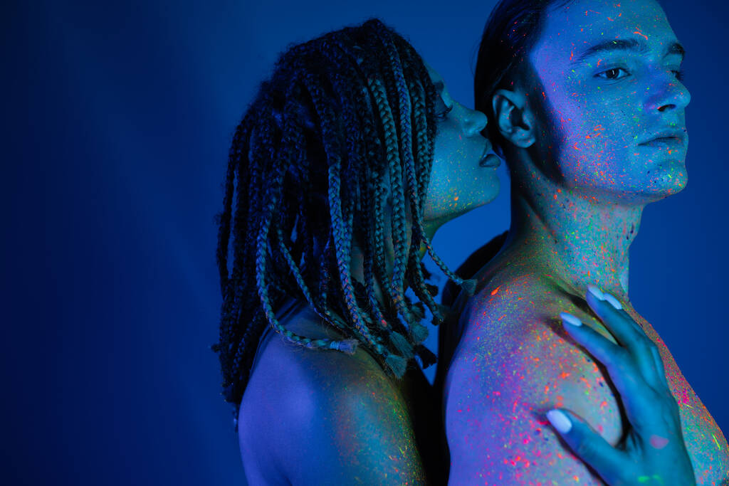 若い異人種間のカップルの親密な瞬間にカラフルなネオンボディペイントで青の背景にシアン照明,印象的なアフリカ系アメリカ人女性抱擁カリスマ男と裸の肩  - 写真・画像