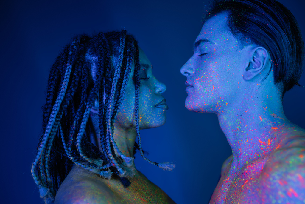 side view of interracial couple with bare shoulders standing face to face with closed eyes, handsome man and african american with dreadlocks on blue background with cyan lighting - Photo, Image