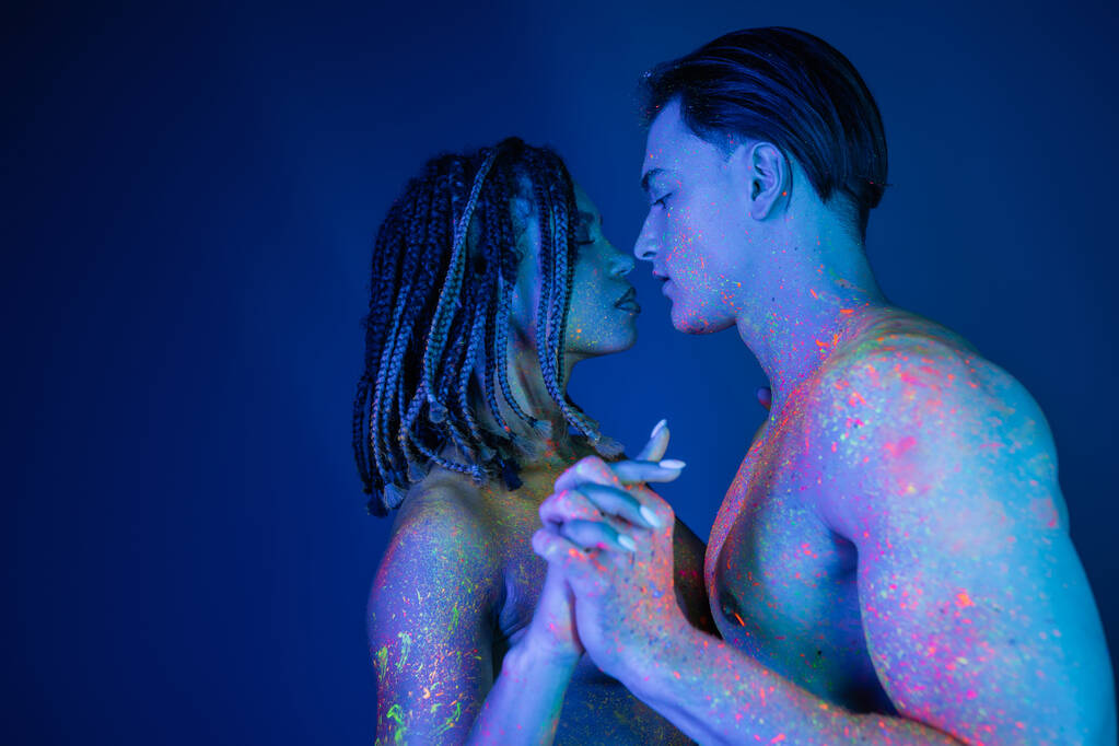 side view of interracial couple in colorful neon body paint standing with clenched hands, african american woman with dreadlocks and shirtless muscular man on blue background with cyan lighting - Photo, Image