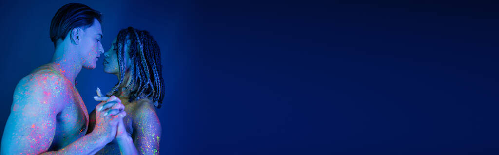 без рубашки мускулистый мужчина и африканская американка с дредами глядя друг на друга и стоя с сжатыми руками в красочной неоновой краске тела на синем фоне с голубым освещением, баннер - Фото, изображение