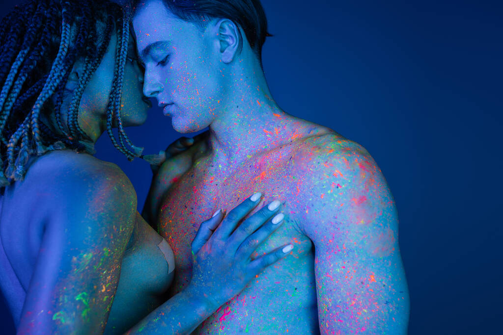 οικεία στιγμή παθιασμένου πολυπολιτισμικού ζευγαριού σε μπλε φόντο με κυανό φωτισμό, γυμνή αφροαμερικανίδα γυναίκα αγγίζοντας το στήθος του γυμνού άνδρα με μυϊκό κορμό - Φωτογραφία, εικόνα