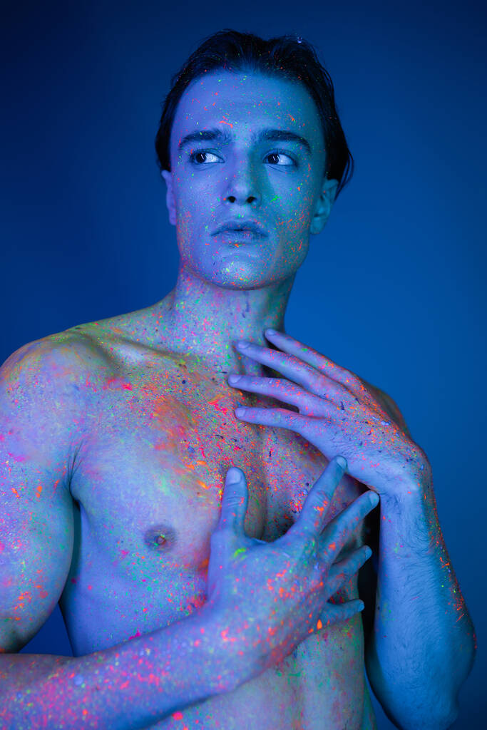 молодий, без сорочки і привабливий чоловік позує в яскравій барвистій неоновій фарбі для тіла, торкаючись голих грудей і дивлячись на синій фон з блакитним ефектом освітлення
 - Фото, зображення
