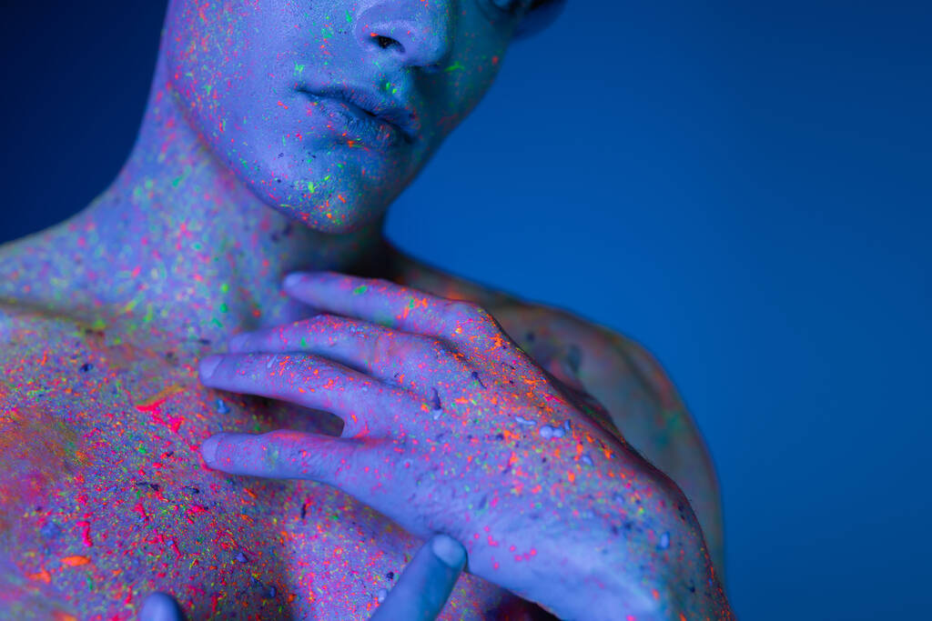 vista recortada del hombre sin camisa cogido de la mano cerca del pecho desnudo mientras posa en colorida y vibrante pintura de cuerpo de neón sobre fondo borroso y azul con efecto de iluminación cian - Foto, imagen