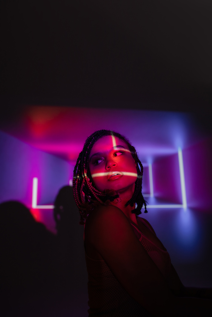 retrato de mujer afroamericana joven y apasionada con rastas mirando hacia otro lado mientras está de pie sobre un fondo abstracto negro y púrpura con rayos de neón y efectos de iluminación - Foto, imagen
