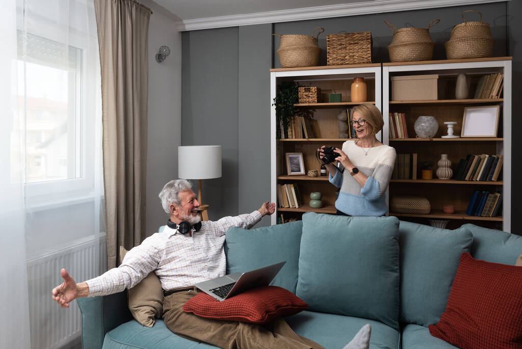Счастливая старшая пара веселится в своем уютном теплом доме. Пожилой мужчина и пожилая женщина просто жить наслаждаясь своей пенсии с книгами, ноутбуком, улыбаясь и говорить о воспоминаниях. - Фото, изображение
