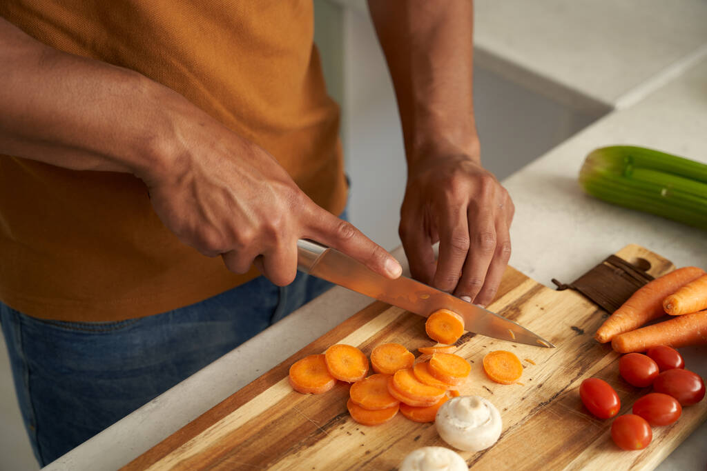Крупный план молодого расового человека в повседневной одежде, нарезающего овощи на кухонной доске - Фото, изображение