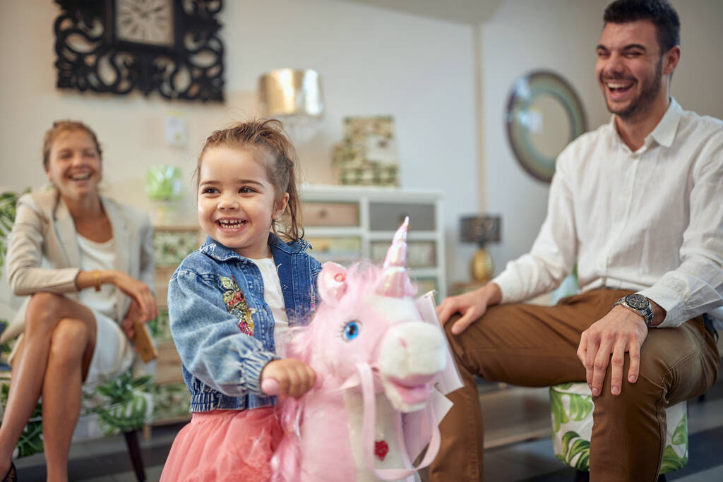 Menina adorável criança montando um cavalo de balanço unicórnio rosa em uma loja de fruniture com seus pais sentados ao lado dela, rindo de alegria, sentindo-se feliz. Compras, família, conceito de estilo de vida. - Foto, Imagem