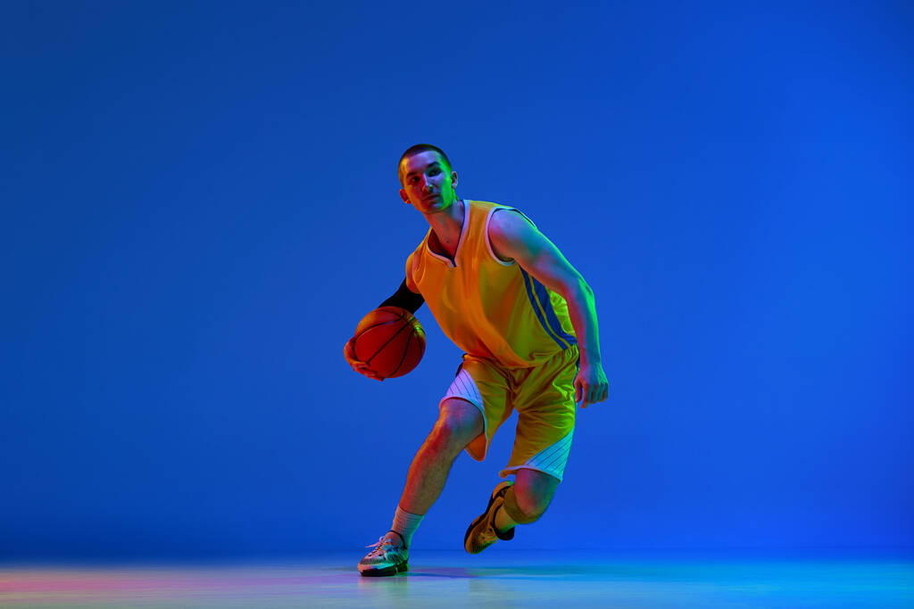 Ανδρική μπασκετμπολίστας με κίτρινη στολή κατά τη διάρκεια του παιχνιδιού, ντρίμπλα μπάλα κατά μπλε φόντο στούντιο σε νέον φως. Έννοια του επαγγελματικού αθλητισμού, χόμπι, υγιεινό τρόπο ζωής, δράση και κίνηση, παιχνίδι - Φωτογραφία, εικόνα