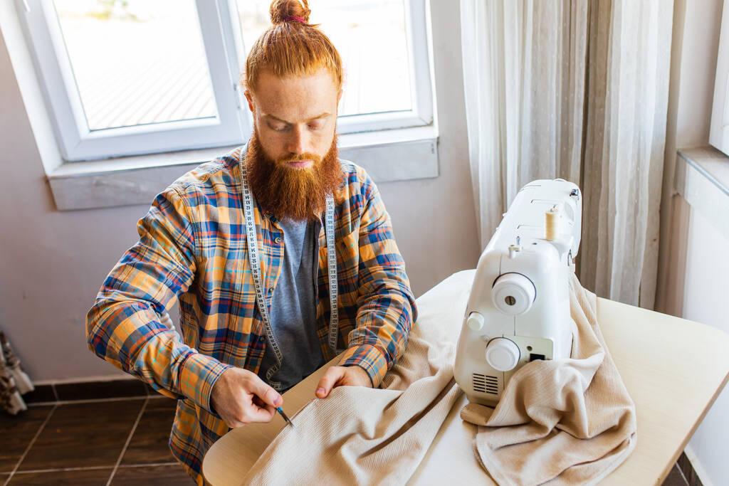 красивый рыжеволосый мужчина с длинной бородой шьет на швейной машинке в домашней студии. - Фото, изображение
