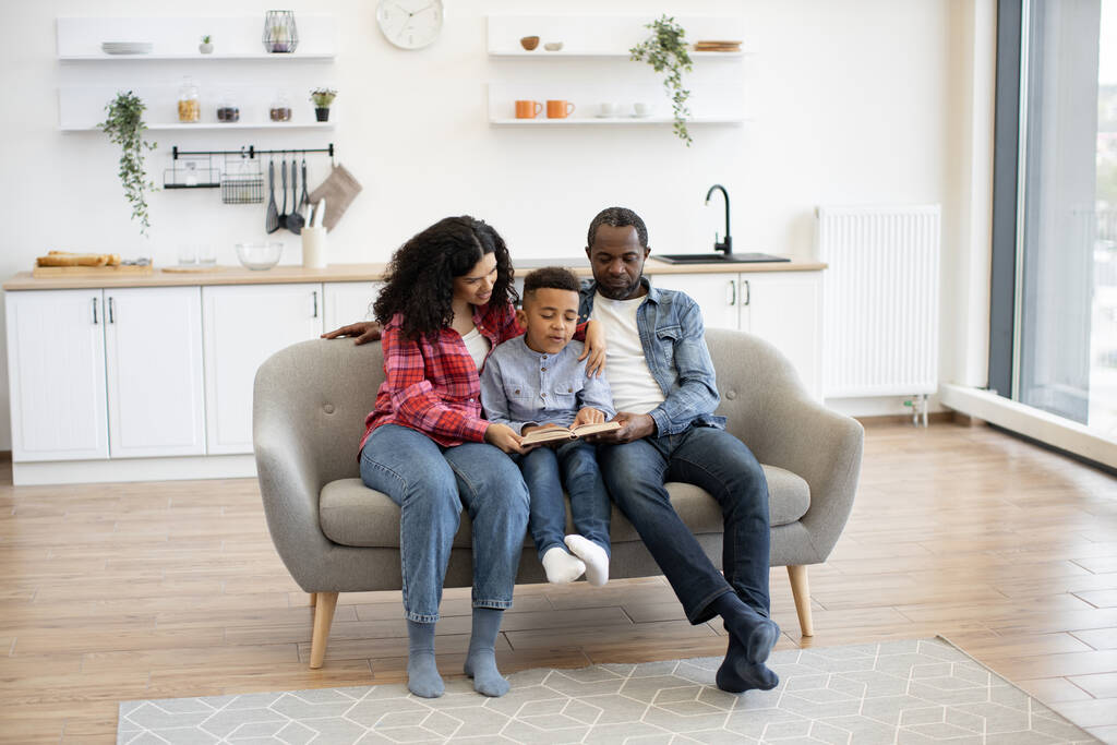Afroamerikanische Ehepartner und kleiner Junge sitzen auf der Couch in der offenen Küche, während sie an einem sonnigen Tag ein Buch in der Hand halten. Nachdenkliche Eltern und Sohn teilen unterhaltsame Aktivitäten beim gemeinsamen Erzählen zu Hause. - Foto, Bild