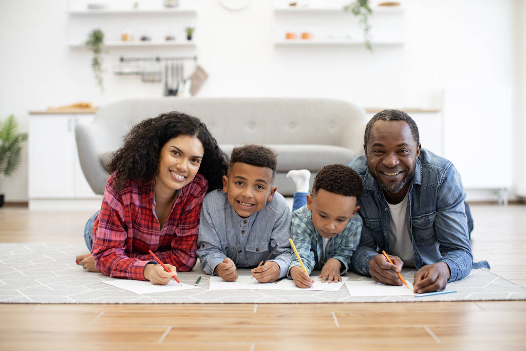 Retrato de familia multiétnica alegre de cuatro lápices multicolores mientras se relaja en la alfombra con sofá detrás. Cónyuges felices y chicos divertidos disfrutando de pasatiempo creativo en la tarde soleada. - Foto, imagen