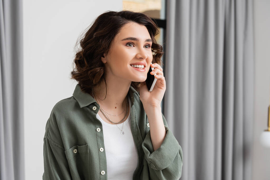τηλεφωνική κλήση, χαρούμενη νεαρή γυναίκα με καστανά μαλλιά φορώντας κομψό πουκάμισο και χαμογελώντας κατά τη διάρκεια της συζήτησης στο smartphone στο δωμάτιο του ξενοδοχείου, ταξιδιωτικός τρόπος ζωής, ζεστή ατμόσφαιρα - Φωτογραφία, εικόνα