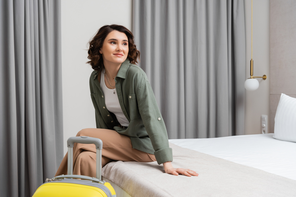 молодая женщина с брюнетками, в повседневной рубашке и брюках, сидит на кровати рядом с желтым чемоданом, смотрит в сторону и улыбается в комфортабельном номере отеля, счастливый путешественник, выходные - Фото, изображение