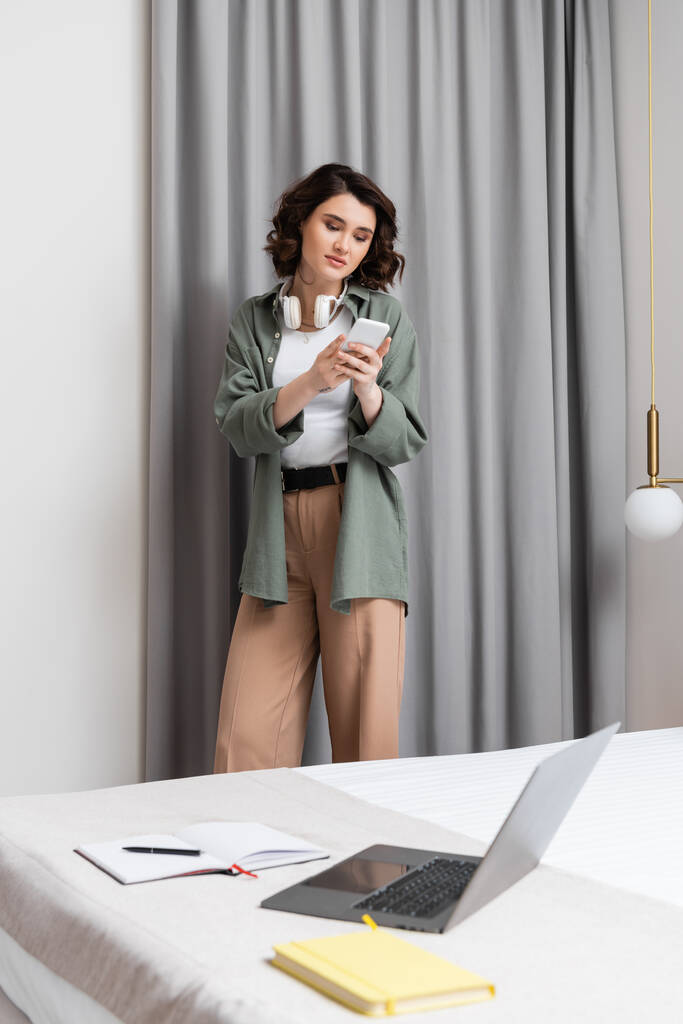 atraktivní žena s bezdrátovými sluchátky stojící v blízkosti šedých záclon, stěnový svícen, postel s laptopem, notebooky a perem, prohlížení internetu na mobilním telefonu v útulné atmosféře hotelového apartmá - Fotografie, Obrázek