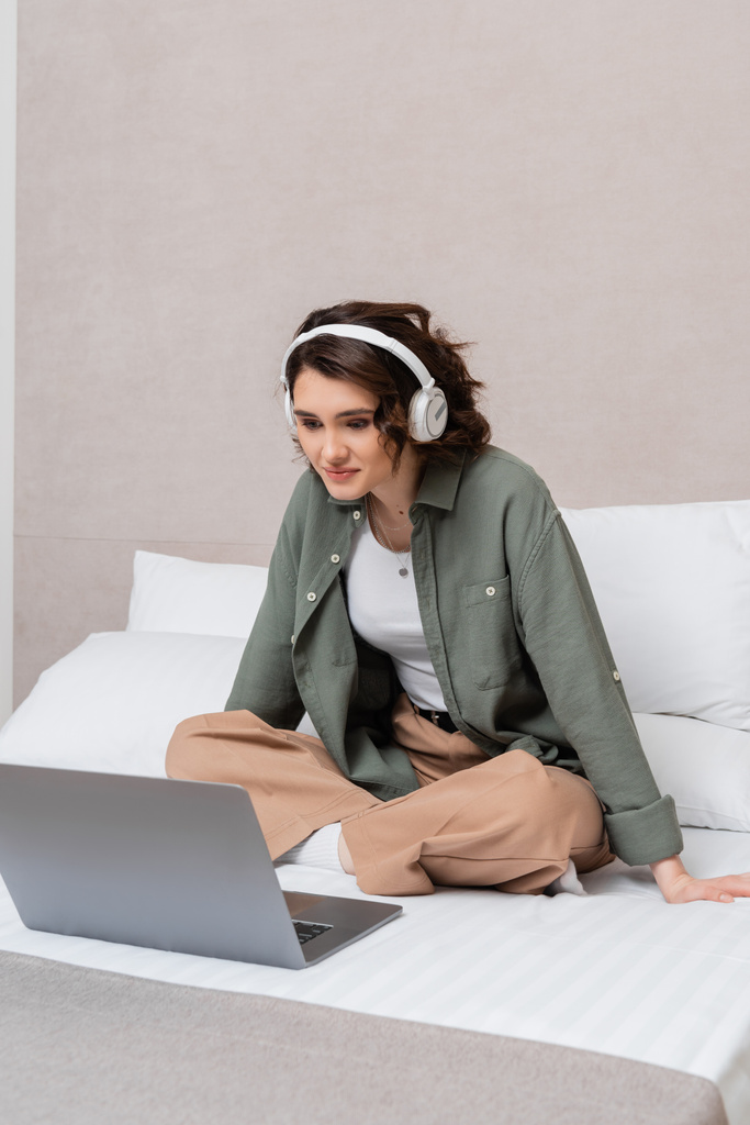 smiling woman with wavy brunette hair, in casual clothes and wireless headphones sitting on bed near white pillows and watching movie on laptop in modern hotel suite, leisure and travel - Photo, Image