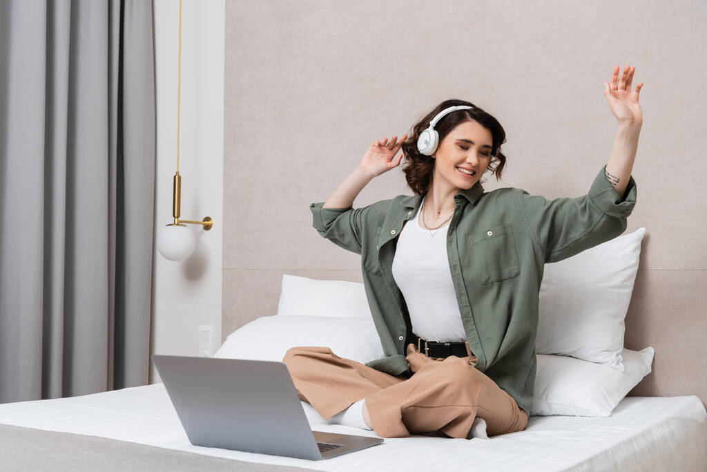 rozradowana wytatuowana kobieta z falującymi brunetkami i zamkniętymi oczami siedząca na łóżku ze skrzyżowanymi nogami w pobliżu laptopa, poduszek i kinkietu, słuchająca podcastu muzycznego i gestykulująca w nowoczesnym pokoju hotelowym - Zdjęcie, obraz