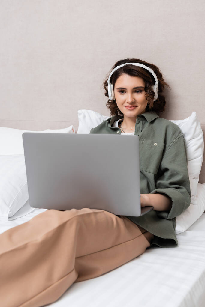 Dalgalı esmer saçlı kaygısız kadın beyaz yastıkların yanında kablosuz kulaklıklar ve günlük kıyafetlerle oturuyor ve otel odasında dizüstü bilgisayardan film izliyor. - Fotoğraf, Görsel