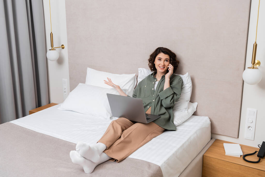 pełna długość radosnych tatuaży kobieta w ubraniach casual siedzi na łóżku z laptopem w pobliżu białych poduszek, kinkiety i szare zasłony, gestykulując podczas rozmowy na telefon komórkowy w nowoczesnym pokoju hotelowym - Zdjęcie, obraz