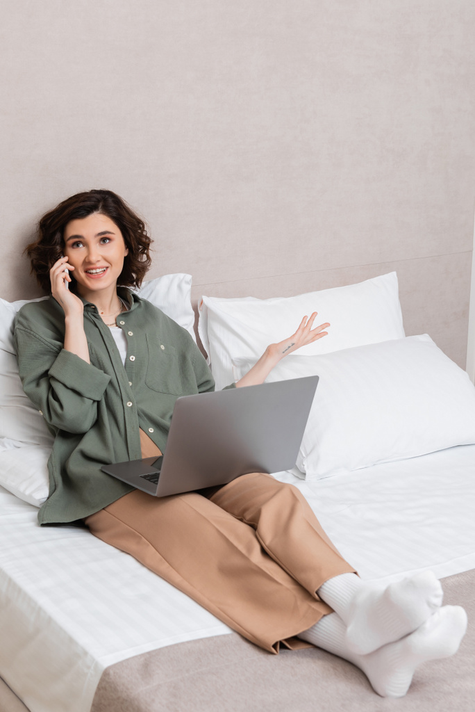 radosna młoda kobieta z falującymi brunetkami i tatuażem, w luźnych ubraniach gestykulująca podczas rozmowy na smartfonie siedząc na łóżku z laptopem w pobliżu białych poduszek w pokoju hotelowym - Zdjęcie, obraz