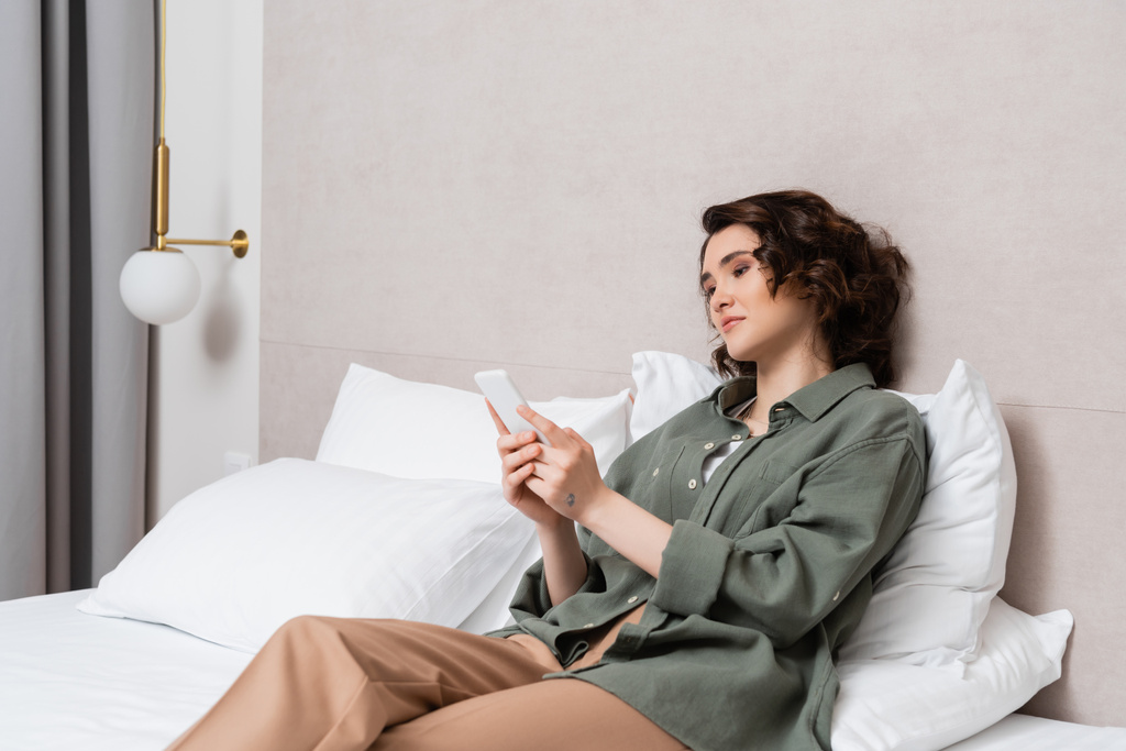 jonge vrouw met golvend brunette haar en tatoeage zitten op bed in casual gesloten en surfen op internet op mobiele telefoon in de buurt van witte kussens en muur sconce in comfortabele hotelkamer, vrije tijd en reizen - Foto, afbeelding