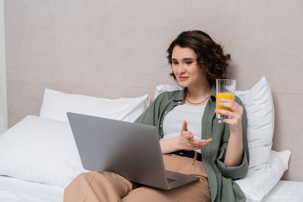 femme souriante aux cheveux bruns ondulés tenant un verre de jus d'orange frais et gesticulant lors d'un appel vidéo sur un ordinateur portable assis sur le lit près d'oreillers blancs et d'un mur gris dans la chambre d'hôtel - Photo, image