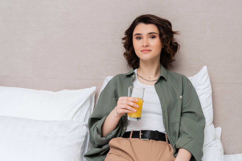 zadowolona kobieta w luźnych ubraniach, z falującymi brunetkami siedzącymi na łóżku ze szklanką świeżego soku pomarańczowego i patrzącymi na aparat w pobliżu białych poduszek i szarej ściany w przytulnym pokoju hotelowym - Zdjęcie, obraz