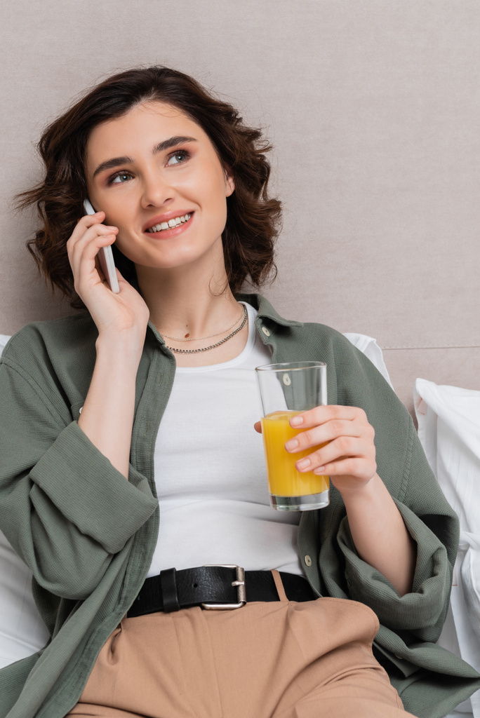 ευτυχισμένη γυναίκα σε casual ρούχα, με κυματιστά καστανά μαλλιά μιλώντας στο κινητό τηλέφωνο στο κρεβάτι κοντά σε λευκά μαξιλάρια και γκρι τοίχο σε ζεστή ατμόσφαιρα της σουίτας του ξενοδοχείου, αναψυχή και ταξίδια - Φωτογραφία, εικόνα