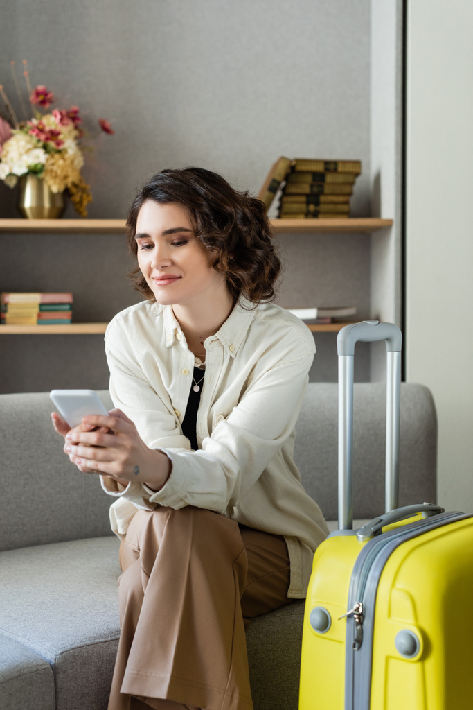 восхитительная татуированная женщина с волнистыми волосами брюнетки сидит на диване рядом с желтой сумкой и просматривает интернет на смартфоне рядом с книгами и вазой с цветами на размытом фоне в лобби отеля - Фото, изображение