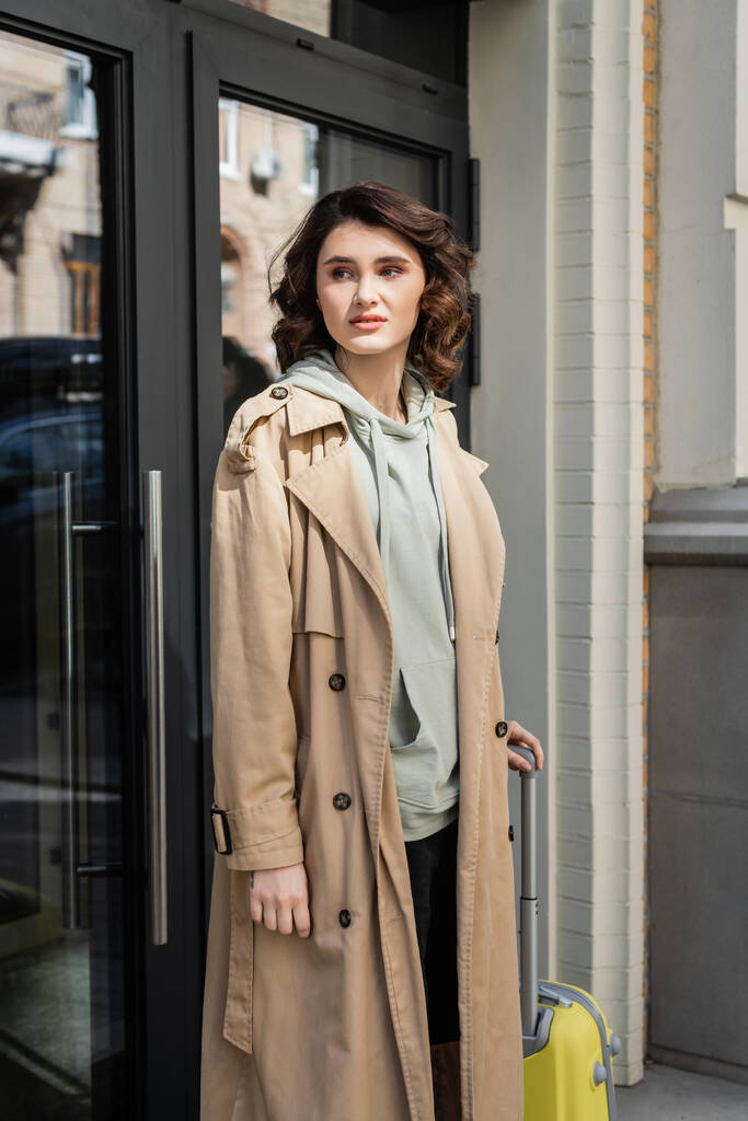 femme attrayante et élégante en trench-coat beige et sweat à capuche gris regardant loin tout en se tenant avec sac de voyage jaune près de l'entrée de l'hôtel moderne sur la rue de la ville européenne - Photo, image