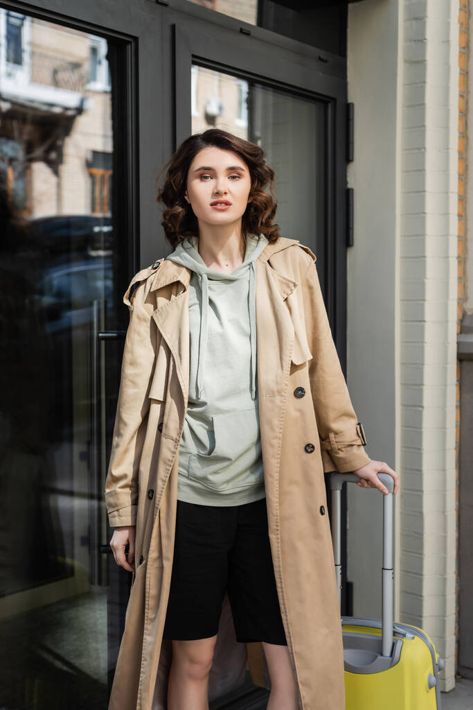 confident woman with wavy brunette hair, in grey hoodie and beige trench coat standing with yellow suitcase and looking at camera near glass doors of modern hotel on city street, travel lifestyle - Photo, Image