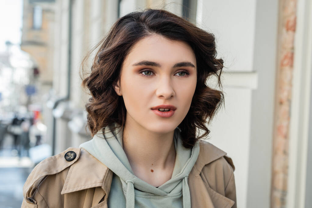 portrait of charming and fashionable woman with short brunette hair, in beige trench coat and grey hoodie looking away in European city on blurred background, street photography - Photo, Image