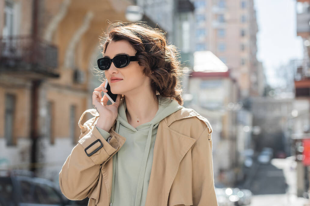 divatos tetovált nő sötét napszemüvegben, szürke kapucnis kapucnis és bézs ballonkabát néz félre beszélgetés közben mobiltelefon a városban homályos háttér, utcai fotózás - Fotó, kép