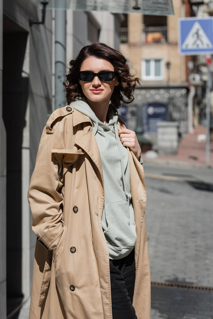 μαγευτική και μοντέρνα γυναίκα με σκούρα γυαλιά ηλίου, γκρι κουκούλα και μπεζ καμπαρντίνα στέκεται στην ευρωπαϊκή πόλη σε θολή φόντο, φωτογραφία του δρόμου, ταξιδιωτικός τρόπος ζωής  - Φωτογραφία, εικόνα