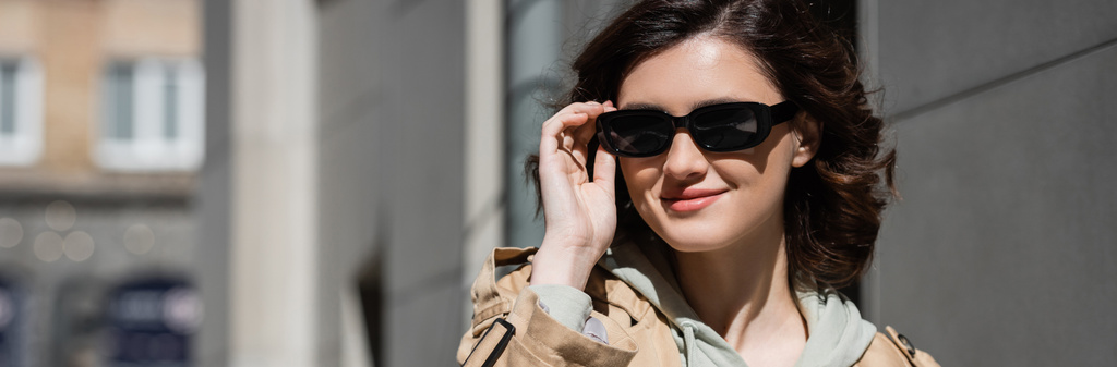 portrait of young and joyful woman with wavy brunette hair, wearing beige trench coat and grey hoodie, adjusting dark stylish sunglasses and looking away on urban street, banner - Photo, Image