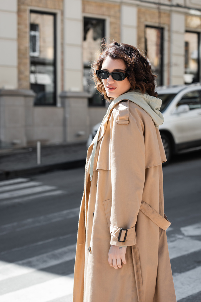 attractive woman with wavy brunette hair walking in dark stylish sunglasses, grey hoodie and beige trench coat while looking at camera on blurred street in European city, urban fashion - Photo, Image