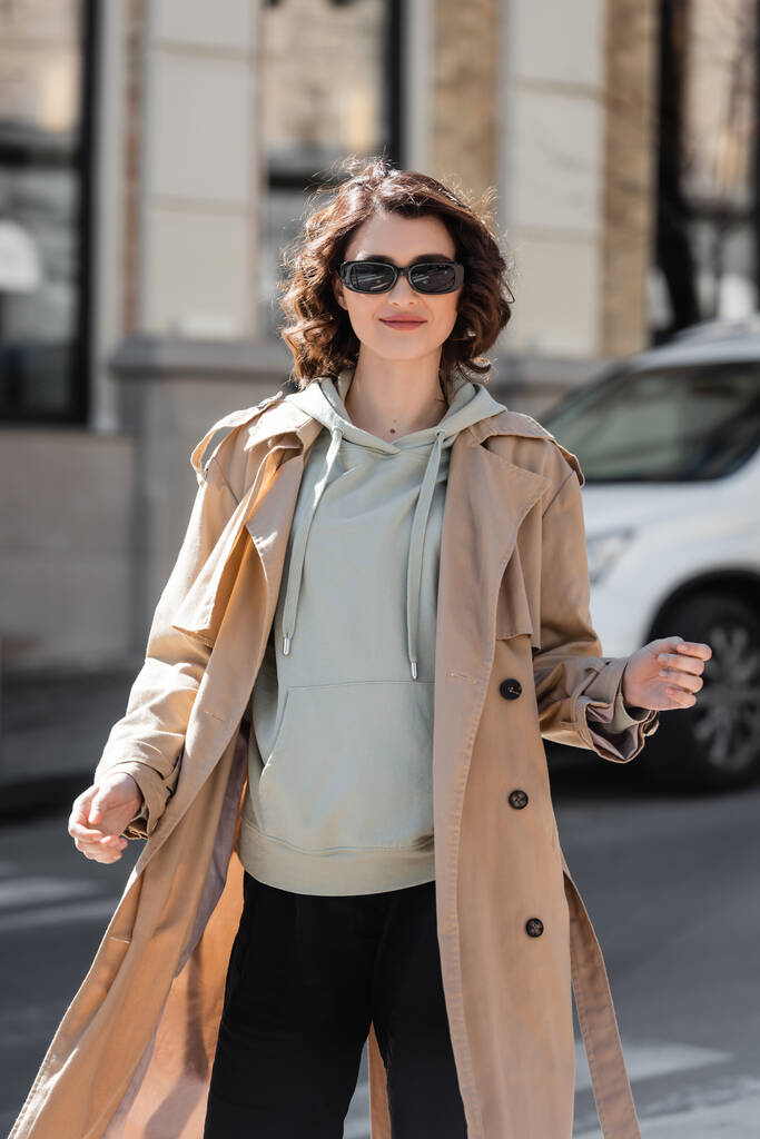 stylische und lächelnde Frau mit dunkler stylischer Sonnenbrille, grauem Kapuzenpulli und beigem Trenchcoat, die in die Kamera schaut, während sie auf verschwommenem Hintergrund durch die Straßen der europäischen Stadt läuft - Foto, Bild