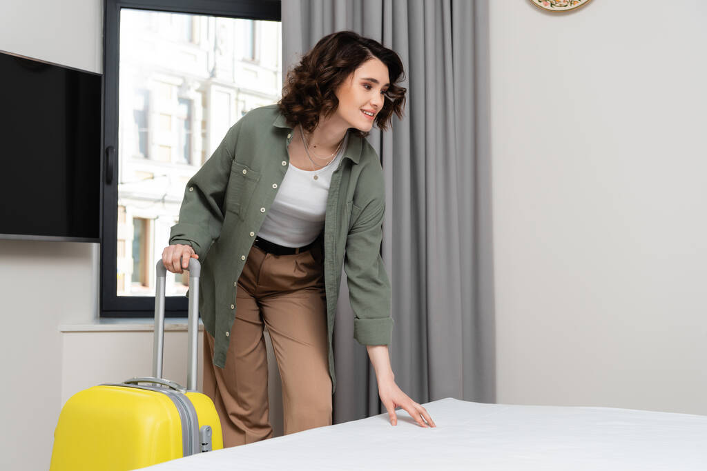 delighted woman in stylish casual clothes, with wavy brunette hair touching comfortable bed in hotel suite near yellow suitcase, window, grey curtains and lcd tv with blank screen, travel lifestyle - Photo, Image