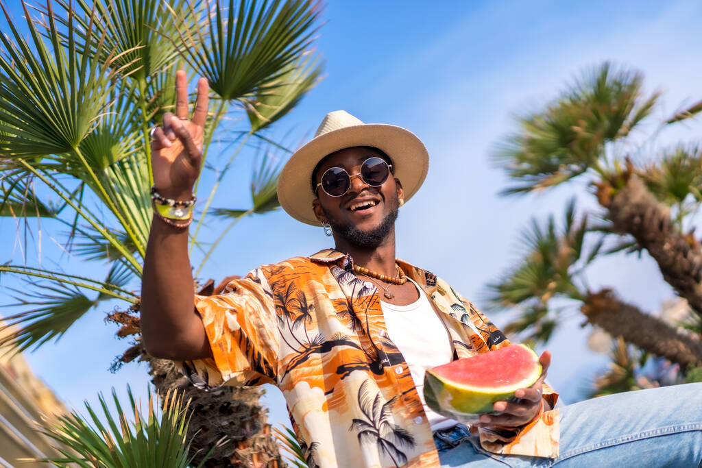 Czarny etniczny mężczyzna cieszący się wakacjami na plaży, w kapeluszu i arbuzie, robi ok - Zdjęcie, obraz